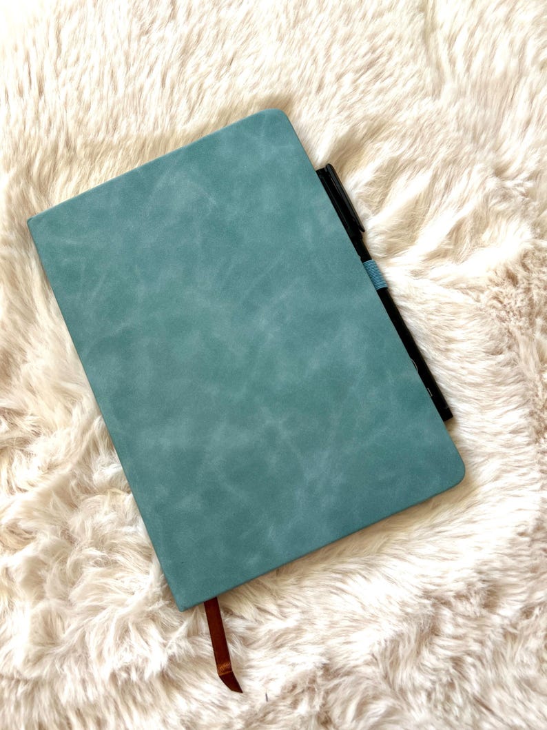 May include: A teal-colored journal with a textured cover and a brown ribbon bookmark. A black pen with a blue band is tucked inside the journal. The journal is resting on a fluffy, off-white surface.