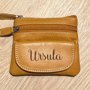 May include: A tan leather coin purse with two zippered compartments and a front pocket. The name "Ursula" is printed on the front pocket in a cursive font. The purse has a light brown stitching.