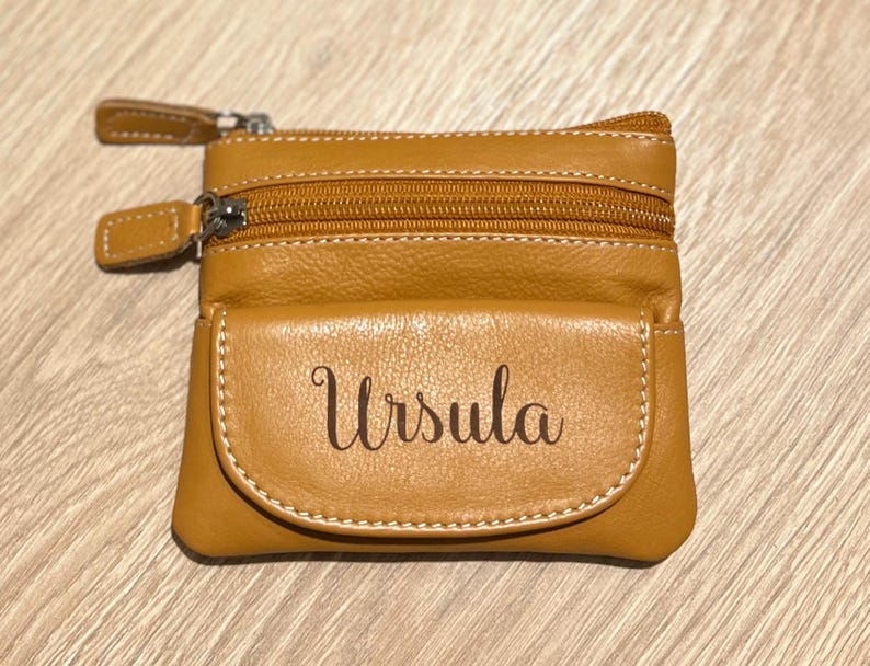 May include: A tan leather coin purse with two zippered compartments and a front pocket. The name "Ursula" is printed on the front pocket in a cursive font. The purse has a light brown stitching.