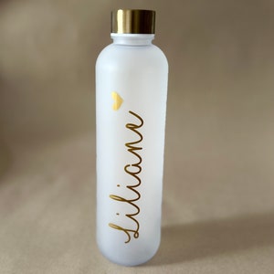May include: A white water bottle with a gold lid and the name "Liliane" in gold script. A small gold heart is above the name. The bottle is frosted and has a sleek, modern design.