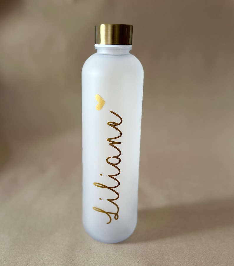 May include: A white water bottle with a gold lid and the name "Liliane" in gold script. A small gold heart is above the name. The bottle is frosted and has a sleek, modern design.