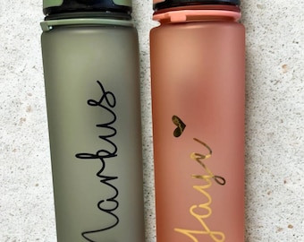 Könnte beinhalten: Zwei zylindrische Wasserflaschen mit mattem Finish. Die linke Flasche ist salbeigrün mit dem Namen Markus in schwarzer Schreibschrift. Die rechte Flasche ist korallenfarben mit einem kleinen Herz und dem Namen Jaye in goldener Schrift.