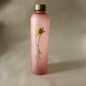 May include: A pink water bottle with a gold-colored lid and a gold floral design. The name "Jaye" is written in a cursive font below the flower. The bottle is cylindrical and appears to be made of glass or a similar material.