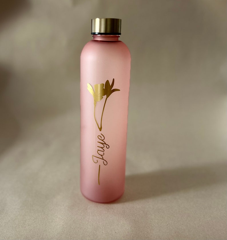 May include: A pink water bottle with a gold-colored lid and a gold floral design. The name "Jaye" is written in a cursive font below the flower. The bottle is cylindrical and appears to be made of glass or a similar material.