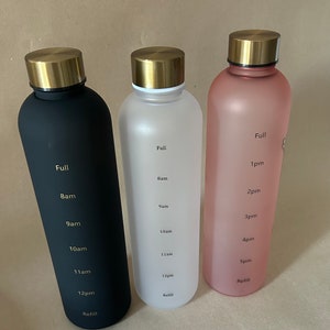 May include: Three water bottles in black, white, and pink, each with a gold-colored lid. The bottles have time markers from "Full" to "Refill" and include times like "8am" and "12pm". The bottles are designed to track water intake throughout the day.