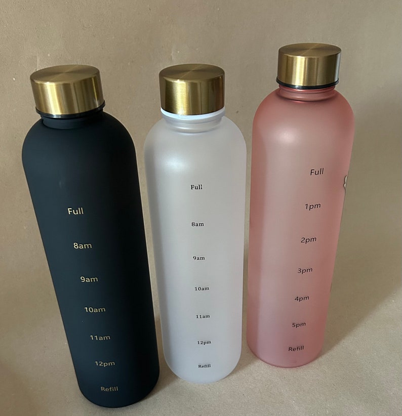 May include: Three water bottles in black, white, and pink, each with a gold-colored lid. The bottles have time markers from "Full" to "Refill" and include times like "8am" and "12pm". The bottles are designed to track water intake throughout the day.
