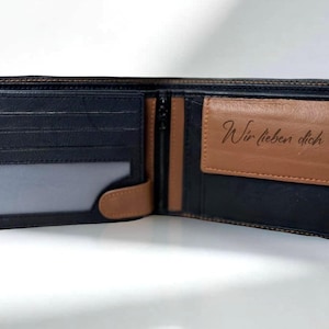 Könnte beinhalten: Geöffnete schwarze Ledergeldbörse mit braunen Akzenten. Die Geldbörse hat mehrere Kartenfächer, ein transparentes Ausweisfach und ein Reißverschlussfach. Ein braunes Lederetikett im Inneren zeigt "Wir lieben dich".