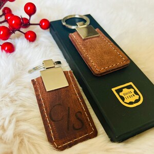 May include: Two brown leather keychains with silver-colored metal accents. One keychain has the initials "C/S" etched into the leather. The keychains are displayed on a white fluffy surface next to a black box.
