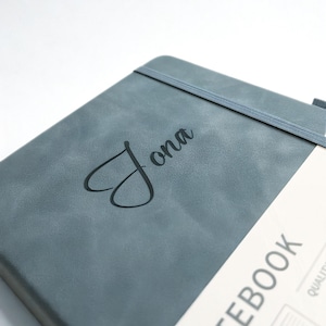 May include: A gray notebook with the name "Jona" in black script and a heart symbol. The notebook has an elastic band closure and the word "NOTEBOOK" printed on the bottom right corner.