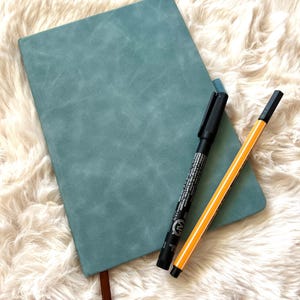 May include: A teal-colored journal with a brown ribbon bookmark and two pens. One pen is black, and the other is yellow with black stripes. The journal is lying on a fluffy, white surface.