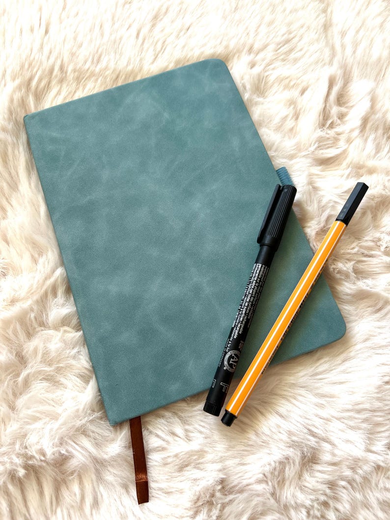 May include: A teal-colored journal with a brown ribbon bookmark and two pens. One pen is black, and the other is yellow with black stripes. The journal is lying on a fluffy, white surface.