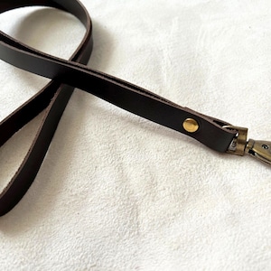 Könnte beinhalten: Ein dunkelbraunes Leder-Lanyard mit einem goldfarbenen Metallverschluss und Niete. Das Lanyard besteht aus einem flachen, dünnen Lederstreifen, der zu einer Schlaufe geformt ist. Der Verschluss ist an einem Ende befestigt.