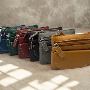 Könnte beinhalten: Eine Auswahl an kleinen Leder-Münzbörsen in verschiedenen Farben, darunter Blau, Grün, Rot, Schwarz, Grau und Braun. Jede Börse hat ein Reißverschluss-Hauptfach, eine Vordertasche und weiße Nähte. Die Börsen sind in einer Reihe angeordnet.