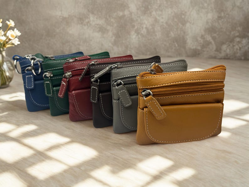 May include: A collection of small leather coin purses in various colors, including blue, green, red, black, gray, and tan. Each purse features a zippered main compartment, a front pocket, and white stitching details. The purses are arranged in a row.