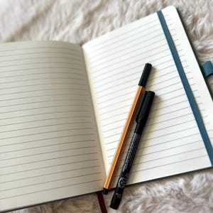 May include: Open notebook with lined pages, a teal elastic band, and a red ribbon bookmark. A black marker and a yellow and black pen rest on the open pages. The notebook is on a white, fluffy surface.