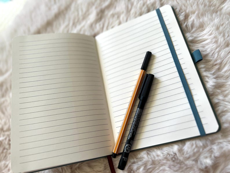 May include: Open notebook with lined pages, a teal elastic band, and a red ribbon bookmark. A black marker and a yellow and black pen rest on the open pages. The notebook is on a white, fluffy surface.