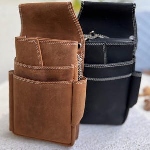 Könnte beinhalten: Zwei Leder-Werkzeuggürtel. Einer ist braun mit weißen Nähten, mit mehreren Taschen und einem Klappenverschluss. Der andere ist schwarz mit weißen Nähten und einem Kettendetail. Beide sind für den Werkzeugtransport konzipiert.