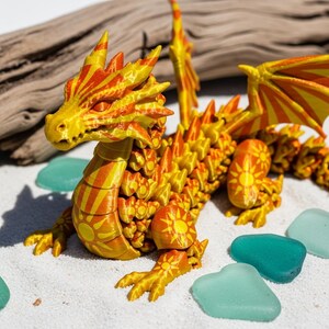 May include: A detailed, 3D-printed dragon figurine in shades of yellow and orange, with a sunburst pattern. The dragon is posed on a sandy surface, with teal-colored sea glass scattered around it. The dragon has detailed wings and a spiky spine.