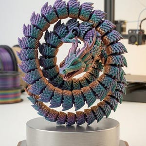May include: A 3D-printed articulated dragon model in shades of purple, teal, and bronze, coiled on a silver and wood base. The dragon has detailed scales and a fierce expression. The base has a label that reads "3D PRINTED ARTICULATED MODEL MODEL NO. Flexi-S1."