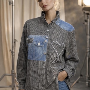 Op de afbeelding: Een grijs en zwart geruit overhemd met denim patches op de schouders en borst. Het overhemd heeft een hoge kraag en knopen. Een wit gestikt hartontwerp staat op de voorkant. Het model draagt een blauwe jeans.