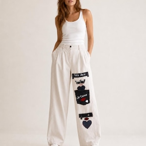 Op de afbeelding: Een wit geribbelde tanktop en een wijde, gebroken witte broek met een reeks donkere stoffen patches op de linkerpijp. De patches bevatten de tekst "TRÈS JOLIE" en "Je t'aime", samen met geborduurde kroon-, hart- en lipmotieven.