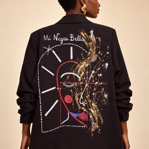 Puede incluir: Un blazer negro con un diseño de rostro abstracto pintado a mano en la espalda. La obra incluye salpicaduras de pintura blanca, roja y dorada, un detalle de labios rojos y el texto cursivo blanco "Mi Negra Bella" sobre la ilustración gráfica.