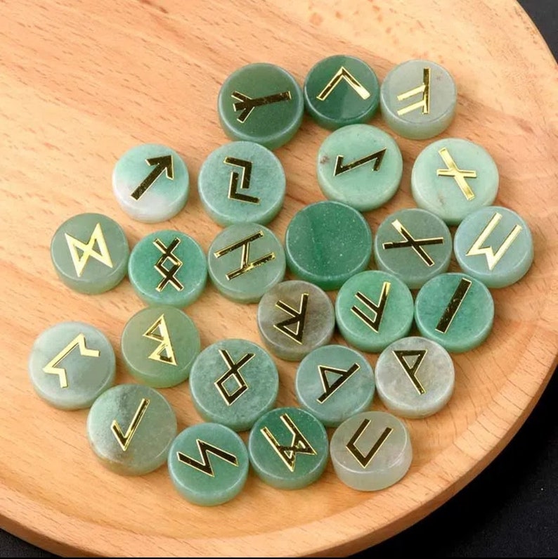 May include: A set of 25 green stone runes with gold lettering. Each rune is a different symbol from the Elder Futhark.