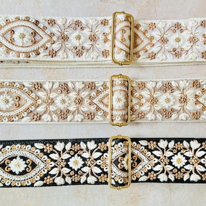 May include: Three decorative bag straps with floral embroidery. The straps are white, beige, and black, with gold-colored hardware. The straps feature an eye-shaped design in the center, surrounded by floral patterns.