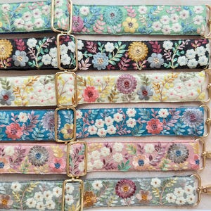 Könnte beinhalten: Eine Auswahl an bestickten Taschenriemen in verschiedenen Farben, darunter Grün, Schwarz, Creme, Türkis, Rosa und Grau. Jeder Riemen zeigt ein Blumenmuster mit goldfarbener Metallhardware und Verschlüssen.