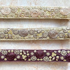 Puede incluir: Primer plano de tres correas decorativas con herrajes dorados. Dos correas son beige con bordados y lentejuelas doradas. La tercera correa es de color púrpura oscuro con bordados y lentejuelas doradas. Las correas son probablemente para un bolso o una bolsa.