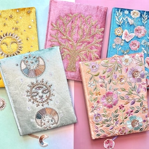 Può includere: Cinque diari decorativi con disegni ricamati. I diari sono in vari colori, tra cui giallo, rosa, blu e pesca. I disegni includono un sole, un albero, fiori, farfalle e motivi celesti. Ogni diario ha un segnalibro a nastro abbinato.