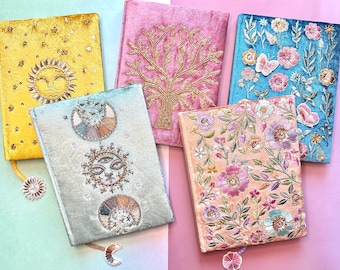 May include: Five decorative journals with embroidered designs. The journals are in various colors, including yellow, pink, blue, and peach. The designs include a sun, a tree, flowers, butterflies, and celestial motifs. Each journal has a matching ribbon bookmark.
