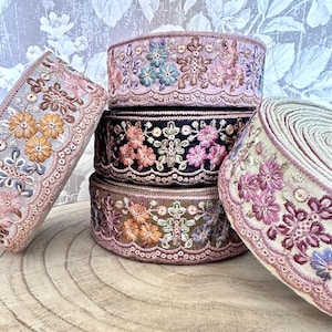 Könnte beinhalten: Eine Auswahl an bestickten Bändern in verschiedenen Farben, darunter Rosa, Schwarz und Beige. Die Bänder weisen florale Muster und aufwendige Details auf, gestapelt auf einer Holzoberfläche. Geeignet für Bastelarbeiten und Verzierungen.