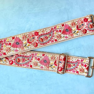 Puede incluir: Una correa de bolso beige con intrincados bordados florales y de cachemira en tonos rosa, rojo, naranja y azul. La correa incluye herrajes de metal dorado, con dos mosquetones giratorios en los extremos y una hebilla deslizante ajustable.