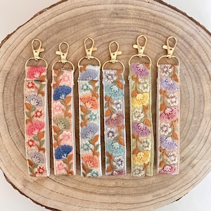 Puede incluir: Seis llaveros coloridos con bordado floral en una correa de tela beige. Cada llavero tiene un cierre de metal dorado.