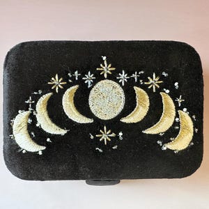 Könnte beinhalten: Schwarze rechteckige Box mit einem gestickten Himmelsdesign. Das Design zeigt einen Vollmond, Halbmonde, Sterne und Perlenakzente in Gold- und Silberfäden. Die Box hat abgerundete Ecken und einen kleinen, schwarzen Verschluss.