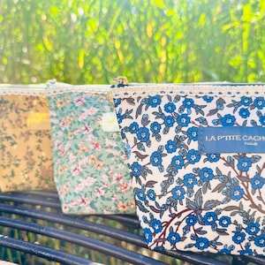 May include: Several floral print pouches in shades of blue, yellow, and green, with lace edging and gold zips. The rightmost pouch has a blue label with the text "LA PTITE CACHOTTIERE PARIS".