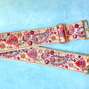 Puede incluir: Una correa de bolso ancha y ajustable con un intrincado patrón de bordado floral y de cachemira en tonos rosa vibrante, rojo intenso y azul oscuro sobre una base de color crema. Incluye herrajes metálicos dorados y ganchos giratorios.