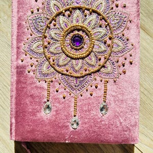 May include: A pink velvet journal with an embroidered mandala design. The mandala features purple and gold thread, a central purple gem, and dangling crystal accents. A matching bookmark is attached.