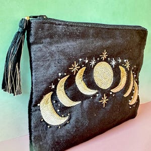 Pode incluir: Bolsa de veludo preta com fases da lua e estrelas bordadas a ouro. O design celestial inclui uma lua cheia e luas crescentes. Um pendão preto com detalhes dourados pende do zíper.