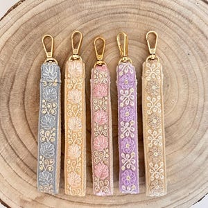 Könnte beinhalten: Fünf bestickte Schlüsselanhänger mit goldenen Akzenten. Die Schlüsselanhänger sind in den Farben Grau, Beige, Rosa, Lavendel und Creme gehalten. Die Schlüsselanhänger werden auf einer Holzoberfläche präsentiert.