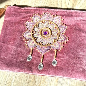 Könnte beinhalten: Eine mauvefarbene Samt-Clutch mit einem gestickten Blumenmuster in Lila- und Goldtönen und einem zentralen lila Edelstein. Die Clutch hat eine goldene Quaste und drei kristallartige Anhänger.
