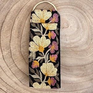 Könnte beinhalten: Ein schwarzer Schlüsselanhänger aus Stoff mit einem goldenen Metallring. Der Stoff ist mit einem floralen Muster in Gelb-, Rosa- und Grüntönen bestickt.