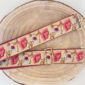 Puede incluir: Una correa ajustable con un patrón floral rojo y beige, con detalles dorados. La correa tiene un diseño floral con acentos rosas, azules y marrones.