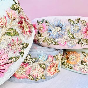Könnte beinhalten: Nahaufnahme von mehreren Rollen floral bestickter Spitzenborte. Die Borte weist eine Vielzahl von Farben auf, darunter Rosa, Blau, Orange und Grün, mit goldenen Akzenten. Die Spitze befindet sich auf einem weißen Mesh-Hintergrund.