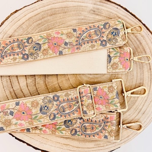 May include: Three beige fabric straps with gold hardware and a paisley and floral pattern. The straps are on a wooden surface.