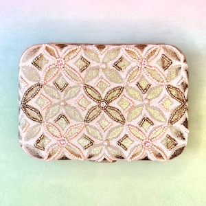 Puede incluir: Clutch rectangular con un estampado floral. El clutch presenta un fondo color crema con diseños florales dorados y marrones. Las flores están adornadas con pequeñas cuentas rosas y perlas. Los bordes del clutch son dorados.