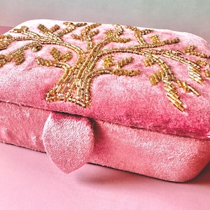 Op de afbeelding: Een roze fluwelen clutch met een gouden kralen boomontwerp. De rechthoekige tas heeft een hartvormige sluiting en een zachte, getextureerde uitstraling. Het boomontwerp is ingewikkeld gedetailleerd met gouden kralen.