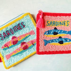 Könnte beinhalten: Zwei quadratische, mit Perlen bestickte Reißverschlusstaschen. Die linke Tasche ist hellblau mit zwei rosa-gelben Fischen und dem Schriftzug SARDINES. Die rechte Tasche ist rosa mit rotem Rand, zwei blau-gelben Fischen und dem Schriftzug SARDINES.