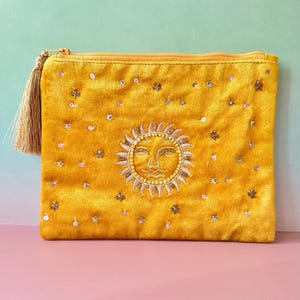 Könnte beinhalten: Senfgelbe Samt-Clutch mit einem Sonnen-Gesicht-Design, das mit goldfarbenem Faden bestickt ist. Die Clutch ist mit Pailletten und kleinen sternförmigen Perlen verziert. Ein goldener Quastenanhänger hängt am Reißverschluss.